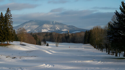 Obraz premium Snowy Golf Course Under a Mountain Range on a Winter Day with Cloudy Skies and Bright Sunlight