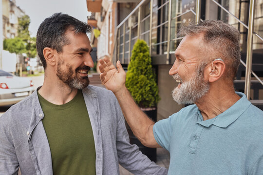 Senior man with hearing impairment uses hearing aid to communicate with his male friend outdoor in noise city. Hearing solutions