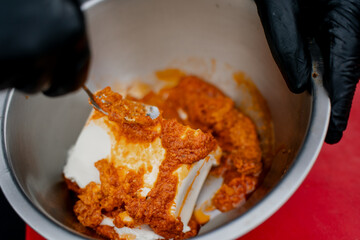 Chef wearing black gloves mixing red spice paste with white creamy ingredient in stainless steel bowl, preparing flavorful marinade or sauce base for restaurant cooking process