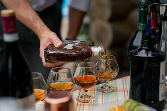 Hand pouring premium cognac from glass bottle into cognac snifters on table with assorted wine bottles, citrus slices and cocktail tools prepared for tasting event