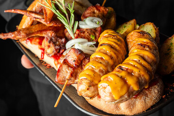 Assorted grilled meats and vegetables on a dark plate, including spiral sausages, skewers, chicken wings, roasted potatoes, onion slices, and rosemary garnish