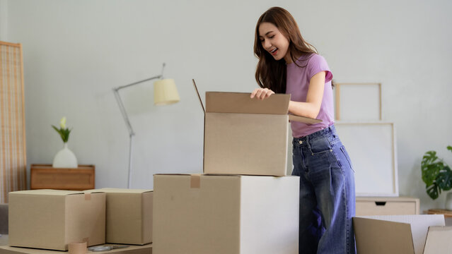 Young Asian Woman Unpacking Moving Boxes in New Home Apartment First Time Homeowner Relocating Content Items from Cardboard Boxes with Smile Enjoy Moving Day