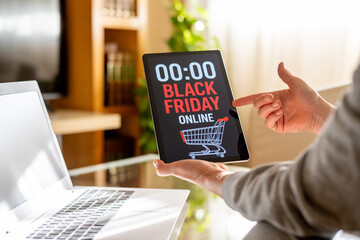 Woman pointing at a digital tablet showing black friday online shopping deals with a shopping cart icon and a midnight timer while using a laptop in her living room