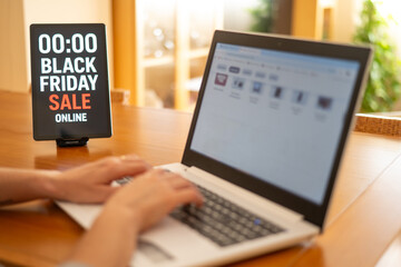 Woman engaging in online shopping during black friday sales, using a laptop while a tablet displays a countdown timer on a wooden table, highlighting the excitement of seasonal deals