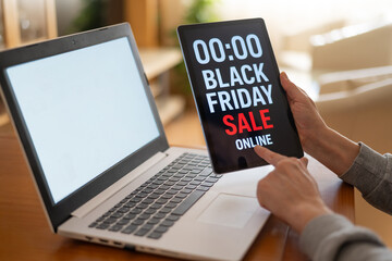 Hands holding tablet displaying black friday online sale announcement next to laptop with blank screen, suggesting online shopping and e-commerce activity