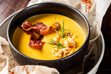 Gourmet pumpkin soup with crispy bacon, rosemary, and popcorn garnish in a black bowl on parchment-lined plate. Rustic autumn dish, top view, ideal for food photography