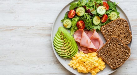 A breakfast plate with scrambled eggs sliced avocado ham mixed green salad and two slices of seeded whole-grain bread on a white wooden table