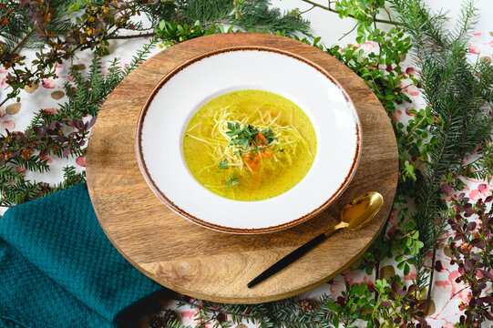 Polish chicken broth (ros&oacute;ł) with thin noodles, fresh herbs and vegetables, served in a white bowl on a rustic wooden board, with a golden spoon and teal napkin, styled with greenery on a light textur