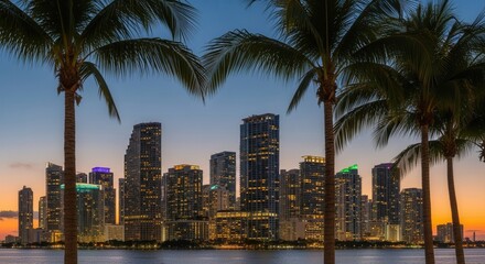 Obraz premium City skyline at dusk with illuminated skyscrapers and palm trees framing a waterfront The sky transitions from blue to orange