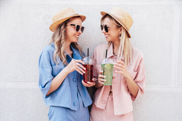 Two young beautiful smiling hipster female in trendy summer clothes. Carefree women posing outdoors. Positive models holding and drinking fresh cocktail smoothie drink in plastic cup with straw