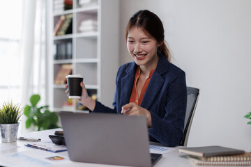 Brunette young asian businesswoman sitting at the office and using phone, laptops for work. Confident professional female wearing business casual
