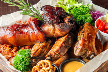 Rustic barbecue platter with glazed pork knuckle, grilled ribs, pulled pork, pretzels, dipping sauce, and fresh vegetables. Hearty meat feast on parchment paper