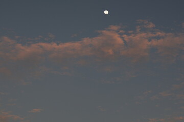 Una toma de cerca de un bello amanecer con un cielo azul y una luna llena con varias nubes de color blanco y gris
