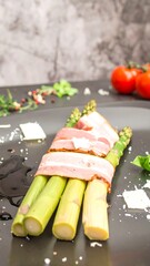 Asparagus spears wrapped in bacon, on dark plate, with cheese, herbs, and tomatoes