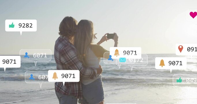 Taking smartphone selfie couple in casual attire hugging at sunset shore, with social media icons
