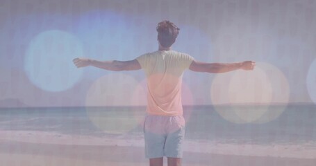 Stretching man wearing pale T-shirt and shorts spreading arms on sandy beach edge facing calm ocean