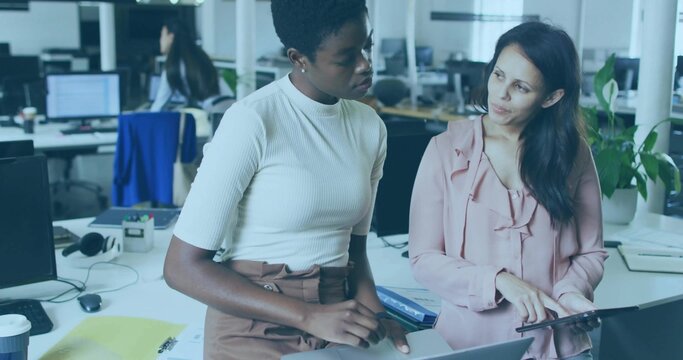 Collaborating women in business attire discussing project at office desk, with laptop and tablet