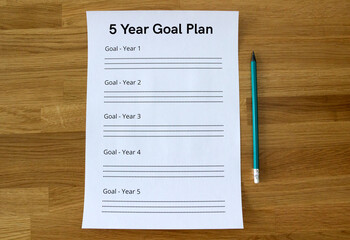 a sheet of paper saying '5 Year Goal Plan' lying straight on the wooden table, with a pencil parallel to the sheet