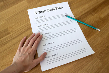 hand holding a sheet of paper saying '5 Year Goal Plan' on a table with a pencil next to it