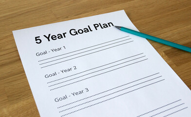 Close up of a paper sheet that says '5 Year Goal Plan' with a pencil pointing at the headline, isolated on wooden background
