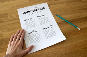 Hand holding a sheet of paper that says 'Habit Tracker' on the wooden table with a pencil next to it, habit tracking concept, healthy lifestyle, sport