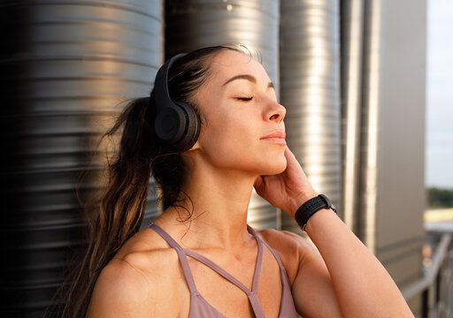 Young female with closed eyes wearing wireless headphones standing at sunset outdoors and enjoying a break during a workout