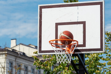 Outdoor basketball hoop with a ball going through the net, set against a clear blue sky and urban buildings in the background © Michele Ursi