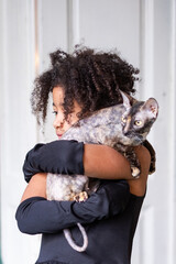 Playful Afro-American girl with curly afro hair embraces her pet cat, showcasing a heartwarming bond in a bright indoor setting with soft textures and warm ambiance