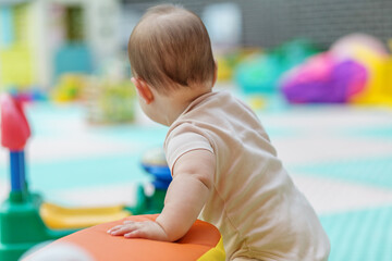 Baby standing in kindergarten playroom with soft toys. Back view