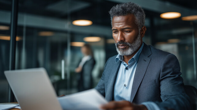 A middle aged Black man works on a laptop a young woman flipping through documents with budget notes their modern office seen through a glass partition top view business