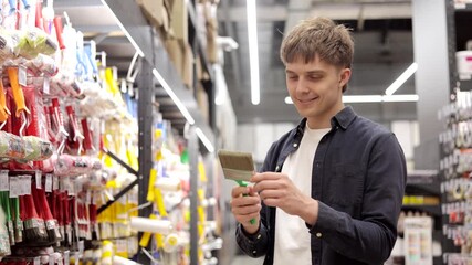 Smiling man standing next to shelf with painting tools choosing paint brush for home renovation in hardware store.