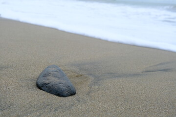 A lone stone rests on smooth sandy beach with ocean waves approaching, symbolizing solitude, calmness, and natural coastal scenery.
