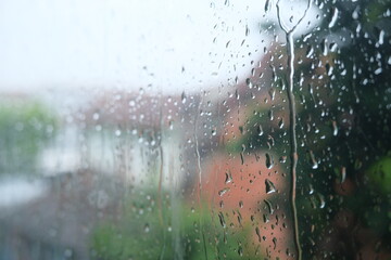 Close-up of raindrops on a glass window with blurred view of houses and greenery in the background.  
