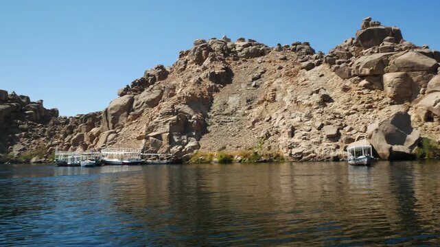 Several traditional touristic boats moored close to an island named "Philae", on the waters of river Nile, Aswan, Egypt. Dry hill on the background, with blue sky.