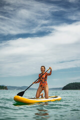 Fit woman in vibrant swimsuit enjoying stand up paddleboarding in turquoise water. Shows energy, balance, and freedom during summer seaside adventure.