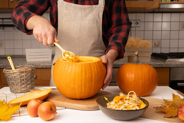 Person in plaid shirt and apron scooping out seeds from a large pumpkin on a wooden cutting board,...