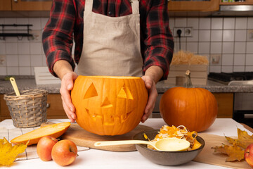 Hands of a person in an apron holding a carved pumpkin with a smiling face, surrounded by autumn leaves, apples, and kitchen utensils, creating a festive atmosphere for Halloween