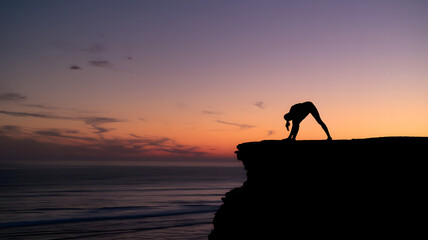 A photo of a silhouette of a person practicing yoga on a cliff during sunrise.