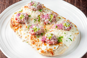 Gourmet flatbread topped with melted mozzarella, raw minced meat, red onion, grated cheese, parsley, and black pepper. Served on a white plate over rustic wooden background.