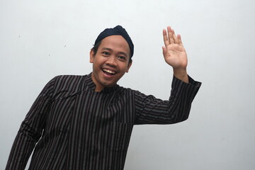 A cheerful man dressed in traditional Javanese attire waves his hand with a big smile, expressing a...