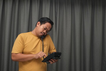 A man in a yellow shirt stands indoors in front of gray curtains, holding a telephone receiver between his head and shoulder while using a tablet with his hands, appearing focused and busy.  
