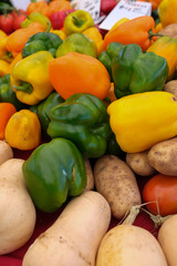 Colorful market display featuring green, yellow, and orange bell peppers alongside light brown potatoes and beige butternut squashes.