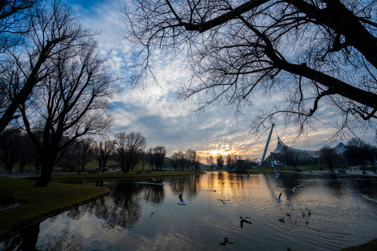 Olympiapark M&uuml;nchen bei Sonnenuntergang mit See, Schw&auml;nen und ikonischer Zeltdacharchitektur