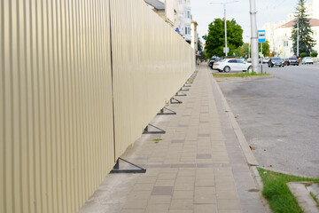 Sidewalk alongside a construction site in an urban area with a long fence and parked cars nearby during daytime