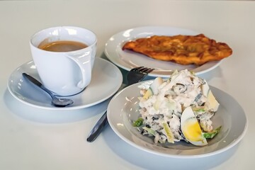 A creamy salad with eggs, a coffee in a white cup, and a fried pastry on a plate present a balanced breakfast trio on a white table