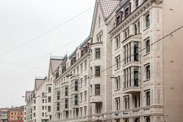 A row of ornate, cream-colored buildings with arched windows and decorative design elements line a city street under a cloudy sky