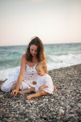 Mother holding baby at the seaside during summer sunset. Happy family moment on the beach with ocean view. Concept of motherhood, childhood, love, and vacation lifestyle outdoors.