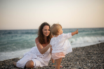 Mother holding baby at the seaside during summer sunset. Happy family moment on the beach with ocean view. Concept of motherhood, childhood, love, and vacation lifestyle outdoors.