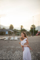 Fototapeta premium Mother holding baby on a rocky beach at sunset. Happy family moment, natural lifestyle, motherhood and childhood concept. Mountains and soft evening light in the background.