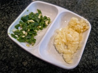 A white divided dish holding chopped green chilies and minced garlic, two common ingredients used for adding spice and flavor in cooking.